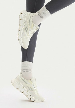 Weiße Reebok-Sneaker, getragen mit weißen Reebok-Socken und dunklen Leggings, ein Fuß angehoben, um die Schuhseite und die Sohlen-Details zu zeigen.