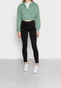 Zkrácená zelená mikina s kapucí a šňůrkami, černé džíny s vysokým pasem a bílé tenisky. Mikina má ribbované manžety a lem.