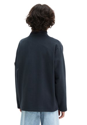 Osoba o kręconych ciemnych włosach, ubrana w czarną kurtkę z długim rękawem i jasnoniebieskie dżinsy, stojąca tyłem na białym tle.