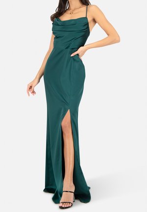Kobieta w długiej do podłogi satynowej sukni w kolorze ciemnej zieleni z cienkimi ramiączkami, rozcięciem z boku i czarnymi, otwartymi na palce szpilkami, pozująca z jedną ręką na biodrze.