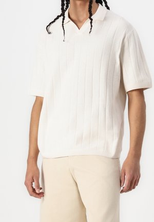 Osoba ubrana w biały, krótki sweter z prążkowanego dzianego materiału z dekoltem w serek, połączony z jasnobeżowymi spodniami.