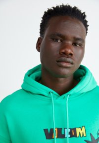 Grön hoodie med dragsko. Har en grafisk text "VOLUME" i svart och gult. Slät tyg och avslappnad passform.