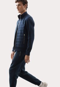 Marineblå lynlåsvest med quiltet front, parret med matchende joggers og hvide sneakers. Har høj krave og lynlåslommer.