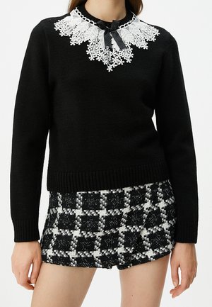Femme portant un pull noir à manches longues avec un col en dentelle blanche et un nœud noir, associé à un short à carreaux noir et blanc.