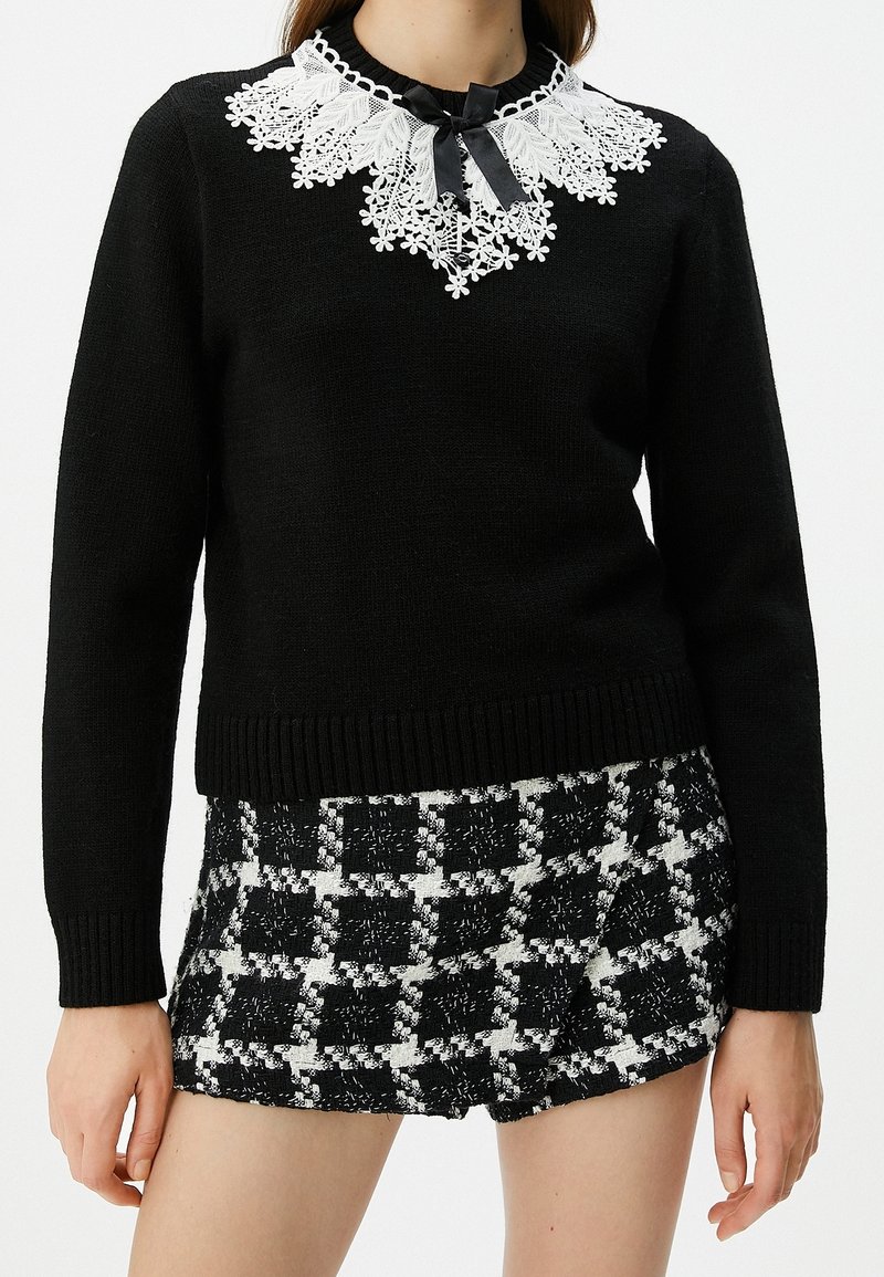 Femme portant un pull noir à manches longues avec un col en dentelle blanche et un nœud noir, associé à un short à carreaux noir et blanc.