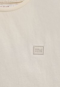 Camiseta de color beige claro con cuello redondo y pequeño logo tonal "POLO CLUB" bordado en el lado izquierdo del pecho. Tejido suave y texturizado.