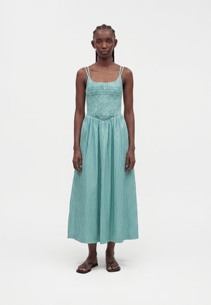 Femme aux cheveux tressés, portant une robe sans manches turquoise smockée à rayures verticales et des sandales noires, debout devant un fond uni.