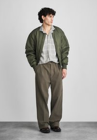 Olive green bomber jacket, gray checkered shirt, and loose brown trousers. Features a zip pocket on the jacket and textured fabric throughout.