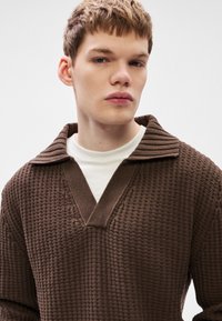 Barna, mintás kivitelű kötött pulóver V-nyakkal és bordázott gallérral. Fehér ing fölé viselve, kiemelve a réteges megjelenést.