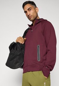 Mann in maroon Zip-Hoodie und grünen Jogginghosen, hält schwarze Sporttasche über der Schulter und posiert vor einem einfarbigen Hintergrund.