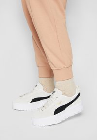 Sneakers bianchi in suede con una striscia laterale nera, dotate di una spessa suola in gomma bianca e branding dorato, abbinate a pantaloni jogger beige e calzini.