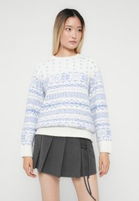 Νεαρή γυναίκα με ίσια ombre μαλλιά, ντυμένη με πουλόβερ λευκού και ανοιχτό μπλε σχεδίου και γκρι πλισέ μίνι φούστα με πλευρικούς κορδόνια.