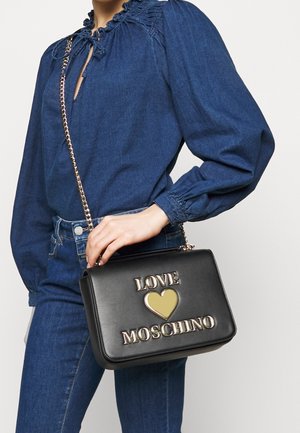 Kobieta w jeansowej bluzce i dżinsach trzyma czarną torebkę Love Moschino z złotym sercem i łańcuszkowym paskiem na wysokości talii.