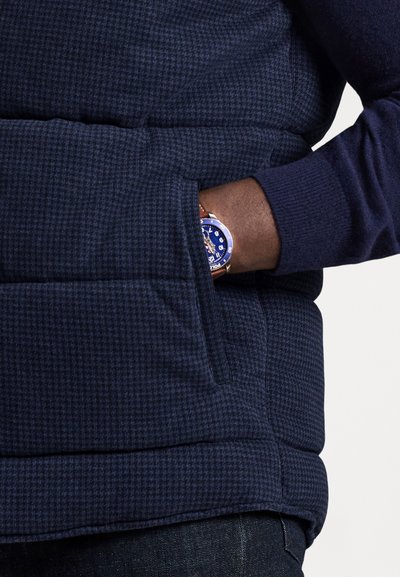 Osoba nosząca granatową, pikowaną kurtkę w kratę, z ręką w bocznej kieszeni, prezentująca zegarek na nadgarstek z niebieską tarczą i brązowym paskiem.