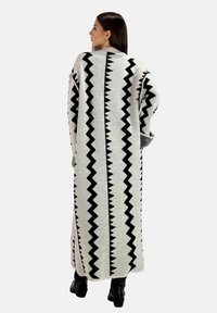 Langer Strickcardigan mit einem Zickzackmuster in Schwarz auf einem hellgrauen Hintergrund. Er hat eine lässige Passform und ein hohes Kragen-Design.