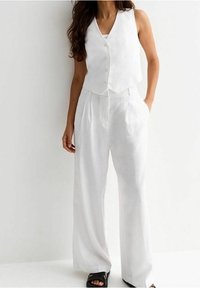 White linen vest with a button front, paired with loose, high-waisted white trousers. Worn with black slide sandals. Smooth texture.