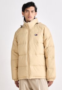 Beige puffer jacket with a quilted design, front zipper, and adjustable hood. Features a small brand logo on the chest. Soft texture.