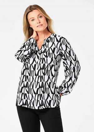 Schwarz-weiß gemusterte Bluse mit langen Ärmeln, Schlagkragen und fließendem Stoff. Kühnes, abstraktes Design im gesamten Kleidungsstück. Kombiniert mit schwarzen Hosen.