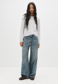 Pulover gri deschis, blugi oversized bleumarin cu un model de stropire, cu formă largă, și încălțăminte cu un model închis, pe un fundal simplu.
