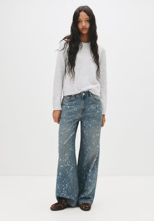 Világosszürke pulóver, túlságosan bő kék denim farmer, fröccsenő mintás, széles lábú fazonnal, és sötét mintás lábbeli egy sima háttér előtt.