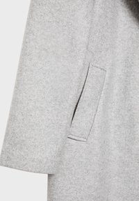 Grå ullrock med mjuk textur, vida ärmar och en diskret ficka längst ner fram, som framhäver en minimalistisk design.