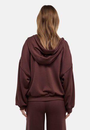 Femme aux longs cheveux brun clair portant un sweat à capuche ample marron foncé et un pantalon assorti, debout de dos sur un fond blanc.