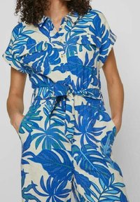 Kombinéza s krátkým rukávem z lehkého materiálu s modro-zeleným tropickým listovým vzorem. Obsahuje dvě náprsní kapsy a detailem uzlu v pase.