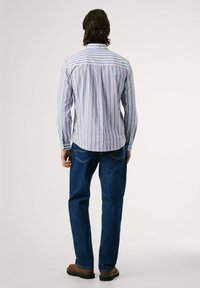 Camisa de manga larga para hombre con rayas verticales azul y naranja. Hecha de tela ligera, cuenta con un yugo trasero y puños con botón. Combinada con jeans azules.