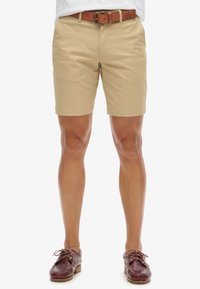 Shorts de algodón beige con frente plano, bolsillos laterales y dobladillo enrollado. Combinados con un cinturón de cuero marrón con una hebilla de metal.