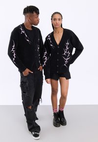 Μαύρες πλεκτές κάρντιγκες με αντιθετικά σχέδια σε σχήμα φλόγας σε μωβ χρώμα. Ο άντρας μοντέλο φοράει σκισμένα μαύρα τζιν, ενώ η γυναίκα μοντέλο φοράει μια μαύρη φούστα.