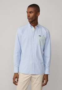 Camicia button-up a righe blu chiaro e bianco con una tasca di un verde uniforme. Presenta un colletto classico e maniche lunghe. Tessuto di cotone.
