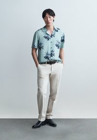 Ung mann med svart hår står med hendene i lommene, iført en lys blå kortermet skjorte med blomster, beige bukser, svart belte og svarte sko.