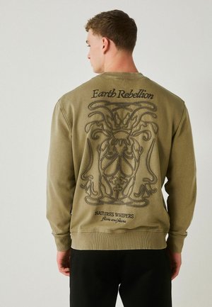 Man wearing olive green sweatshirt with black embroidered abstract floral design and text "Earth Rebellion" and "Natures Whispers flora and fauna" on back.
