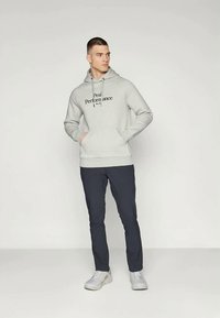 Jeune homme en sweat à capuche gris avec le texte "Peak Performance", en pantalon bleu marine et sneakers blanches, se tenant les mains dans les poches, regardant à gauche.