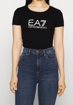 Femme portant un t-shirt noir ajusté avec le logo blanc "EA7 Emporio Armani" et un jean taille haute bleu foncé, debout devant un fond blanc.