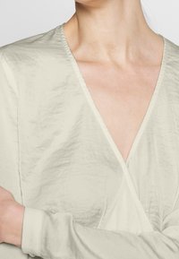 Top avvolgente beige chiaro con scollatura a V. Realizzato in morbido tessuto testurizzato, presenta maniche lunghe e sottili dettagli increspati.