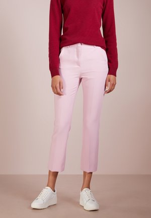 Persona que lleva pantalones cortos de color rosa claro, zapatillas blancas y un suéter de manga larga rojo, de pie sobre un fondo neutro.