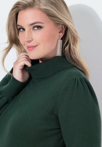 Maglione verde in maglia con colletto alto, dotato di maniche a sbuffo. Orecchini a chandelier scintillanti aggiungono un accento distintivo. Texture liscia.