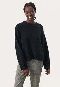 Dunkel gestrickter Pullover mit Rundhalsausschnitt, weiten Ärmeln und geripptem Saum. Kombiniert mit grauen karottenförmigen Hosen.
