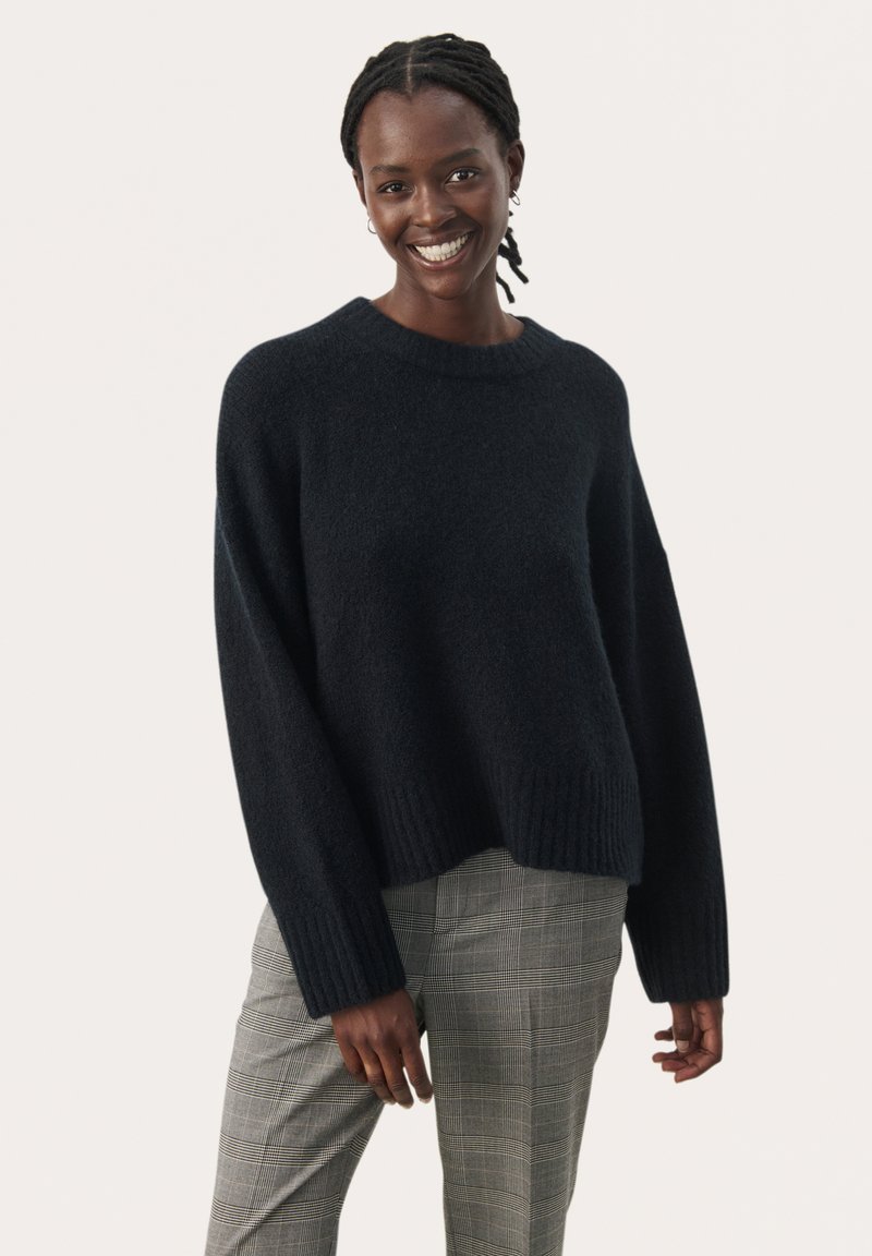 Dunkel gestrickter Pullover mit Rundhalsausschnitt, weiten Ärmeln und geripptem Saum. Kombiniert mit grauen karottenförmigen Hosen.