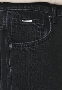 Jeans de mezclilla negra con un bolsillo delantero, etiqueta de marca con "PEGADOR" y una textura sutil con un corte clásico. Acentuados con herrajes de metal.