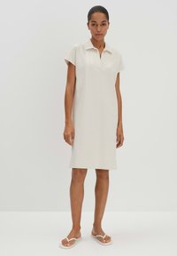 Beiges Polokleid mit kurzen Ärmeln, geradem Schnitt und Kragen. Glatte Textur, knielang, kombiniert mit weißen flachen Sandalen.
