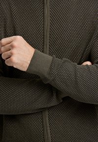 Mörkgrön texturerad zip-up hoodie med ett bikakesmönster. Har ribbade muddar och en slät dragkedja på framsidan för stängning.