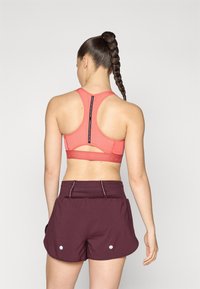 Racerback sports bra in coral with mesh detailing and a cutout; paired with maroon shorts featuring side pockets and accent stitching.