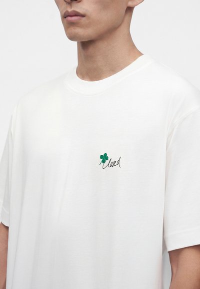 Homme portant un t-shirt blanc uni avec un petit trèfle à quatre feuilles vert et le mot "loved" brodé sur le côté gauche de la poitrine.