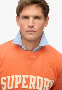 Oranje sweatshirt met 'SUPERDRY' logo in crème. Lichtblauwe kraag zichtbaar eronder. Zachte textuur, casual ontwerp, standaard pasvorm.