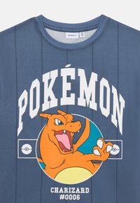 Mėlyna trumpomis rankovėmis marškinėliai pagaminti iš kvėpuojančios medžiagos, su dideliu Charizard grafiniu atvaizdu ir „POKÉMON“ tekstu balta spalva, o apačioje - #0006.