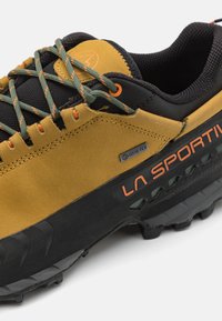 GORE-TEX túracipő barna velúr felsőrésszel, fekete díszítéssel, zöld és narancssárga fűzőkkel, textúrált fekete talppal és "LA SPORTIVA" márkajelzéssel.