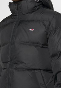 Zwarte pufferjack met hoge kraag, ritssluiting aan de voorkant en geborduurd logo op de linkerkant van de borst. Heeft een gestikte design en een capuchon met trekkoord.