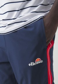 Marina shorts med röda sidränder och en Ellesse-logotyp. Materialet verkar lätt och slätt, med en figurnära design. Vit och marinrandig skjorta ovanför.